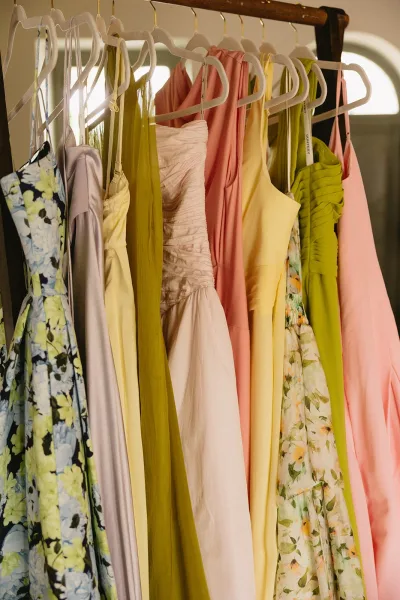 Bridesmaid dresses in a mismatched bridesmaid dress palette hang on a rack, satin and chiffon styles with spaghetti straps in window light