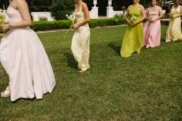 Bridesmaid procession with pastel satin dresses and calla lily bouquets, walking across a grass lawn in a hedge-lined courtyard