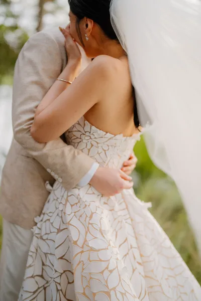 Bride and groom hug in a tender first look moment beneath garden trees, her long veil trailing as he holds her waist.