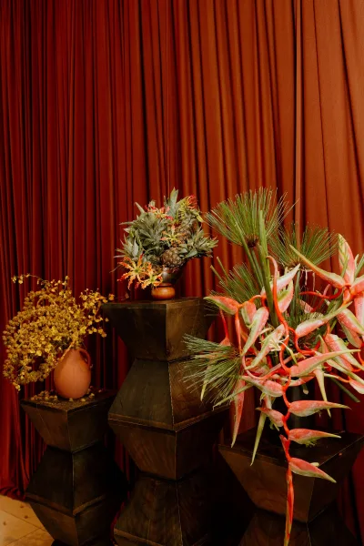Floral installation with pedestal flower arrangement in a terracotta vase, featuring layered, sculptural forms set against rust drapery.