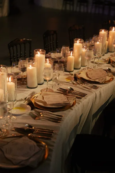 Reception tablescape set as a candlelit wedding table with pillar candles, in a dim reception hall with black chairs.