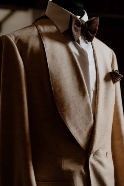 Groom suit, a tan wedding suit accented by a brown bow tie, crisp tailoring shown against a dark indoor backdrop