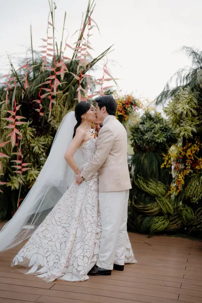 Wedding kiss portrait as the bride and groom embrace beneath a tropical ceremony floral installation, her long tulle veil catching light.