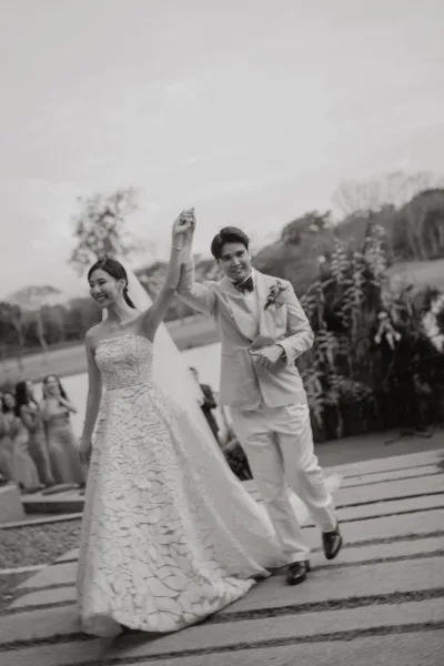 Wedding recessional with bride and groom walking hand in hand, her veil trailing, along a stone walkway beneath garden trees by the lake.