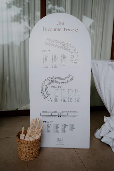 Wedding seating chart on an arched wedding sign, flanked by paper fans, set by a glass door with white curtains and warm wood trim.