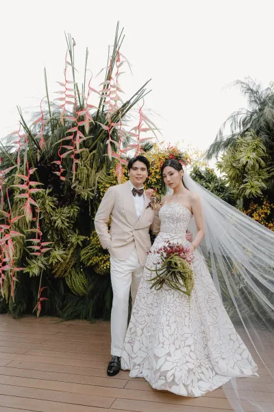 Couple portrait of the bride and groom standing, the bride holding a bouquet, framed by tropical foliage and hanging flowers.