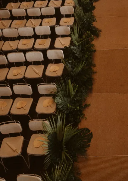 Ceremony aisle decor with greenery aisle runner and tropical leaves lining a wood floor aisle beside folding chairs with programs and fans