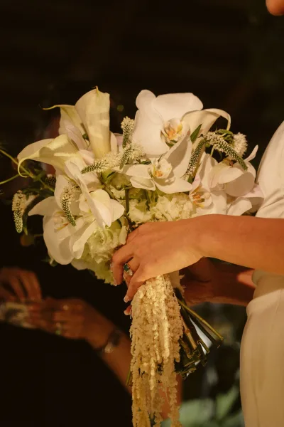 Bridal bouquet of white orchids and calla lilies cascading with greenery, held by bride’s hand with engagement ring in dark indoor setting