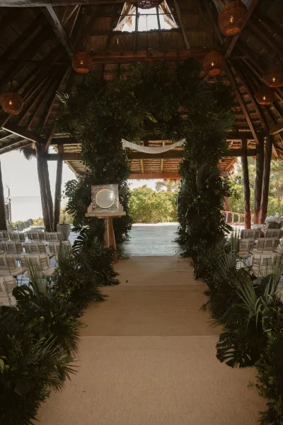 Ceremony aisle decor with tropical wedding ceremony greenery arch, palm fronds and monstera lining a runner in a thatched pavilion by the ocean.