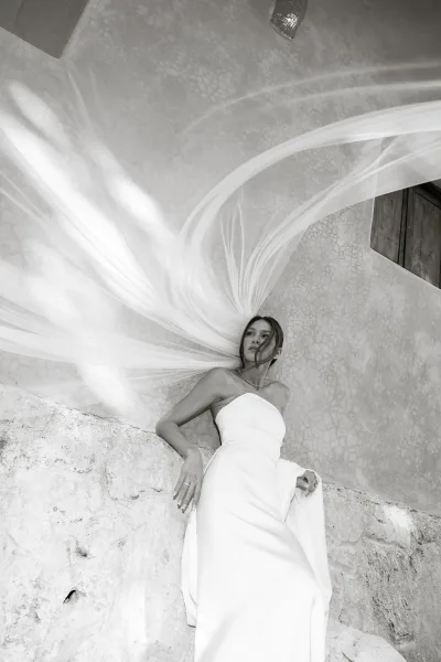 Bridal portrait in black and white of a bride in a strapless wedding dress with long veil, leaning by a stucco wall with wooden shutter