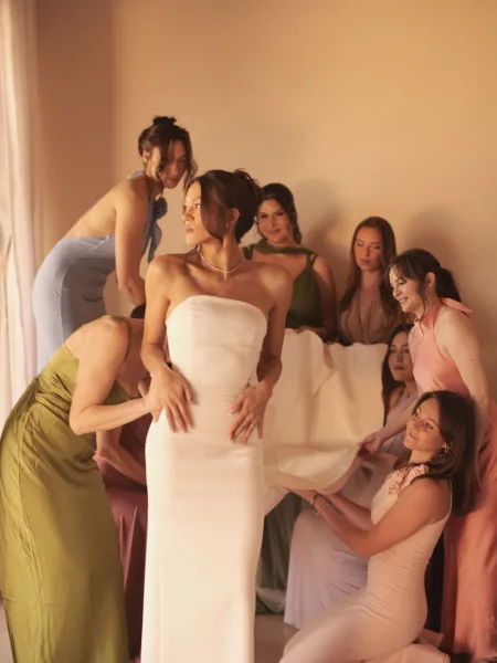 Bridesmaid group photo of the bride with bridesmaids in satin pastel dresses, standing by a neutral wall in soft window light