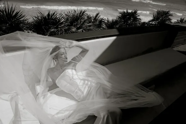 Bridal portrait of a bride with veil draped over her face, reclining on concrete stairs by ocean waves and palm tree shadows