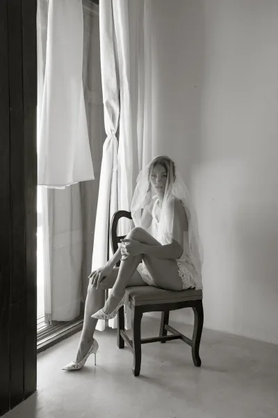 Bridal portrait of a bride in lace lingerie and heels, veil draped over her face as she sits by a window with sheer curtains