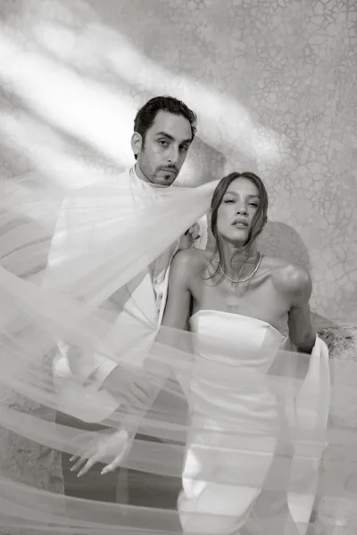 Couple portrait with a wedding veil draped over them, bride in strapless dress and pearls beside groom in white suit on stone steps