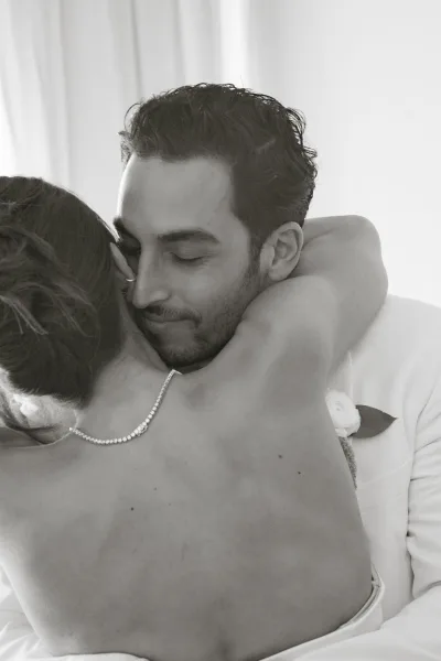 Wedding couple embrace in soft indoor light, bride hugging groom in a white suit with boutonniere before white curtains