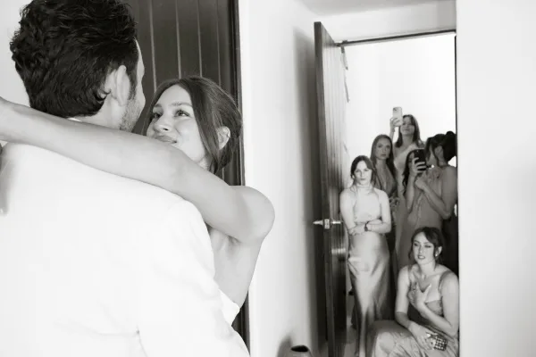 First dance as bride in a strapless wedding dress hugs groom in a white suit while bridesmaids watch and a phone snaps photos indoors