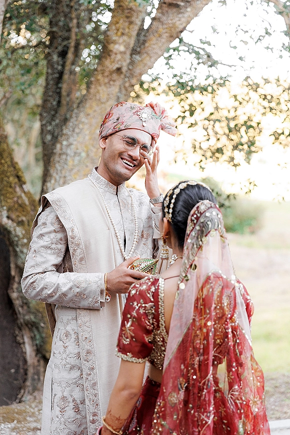 First look moment as groom in embroidered sherwani wipes tears facing bride in red lehenga on a sunlit outdoor path by greenery