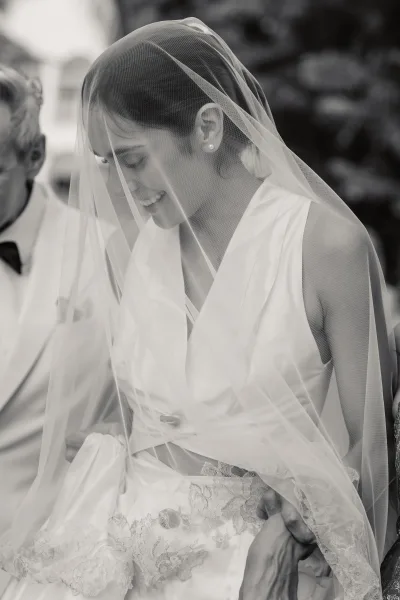 Bridal portrait of a bride under veil, softly framed by sheer tulle, with trees and a building facade subtly blurred in the background.