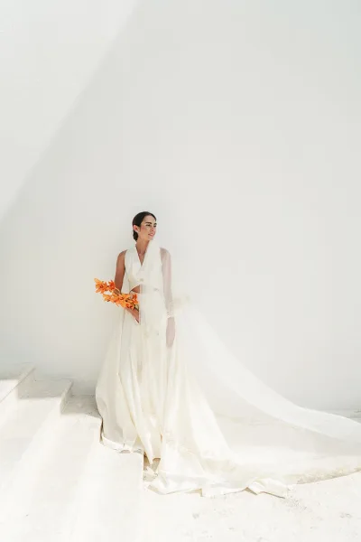 Bridal portrait of a bride with bouquet on stone steps by a white wall, highlighting an orange orchid bouquet against a clean backdrop.