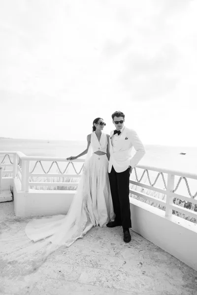 Couple portrait of a bride in a halter neckline wedding dress and a groom in a white dinner jacket, wearing sunglasses on an oceanfront terrace.