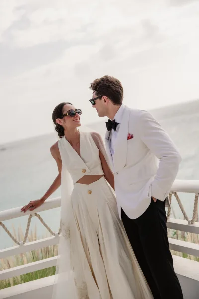 Bride and groom portrait featuring modern bridal separates, both in sunglasses, on a seaside balcony overlooking ocean, sky, and coastal grasses.