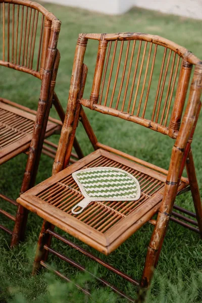 Ceremony chairs styled as rattan wedding chairs, each seat holding a woven hand fan, arranged in tidy rows for arriving guests.