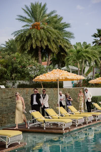 Wedding guests mingle as poolside wedding guests beneath palm trees, one in a white dinner jacket, beside a turquoise swimming pool and stone wall.