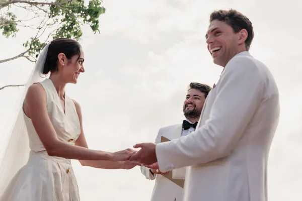 Ceremony moment of the couple exchanging wedding vows, the groom in a white tuxedo, under open sky with tree branches framing their smiles.