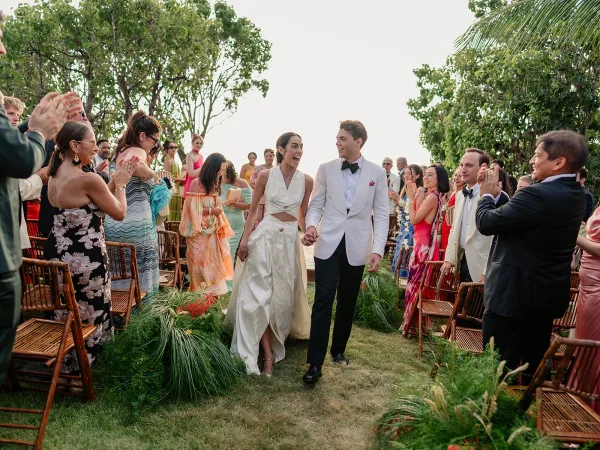 Ceremony exit with a couple holding hands as guests cheer along a garden aisle lined with lush aisle greenery beneath swaying palm fronds.