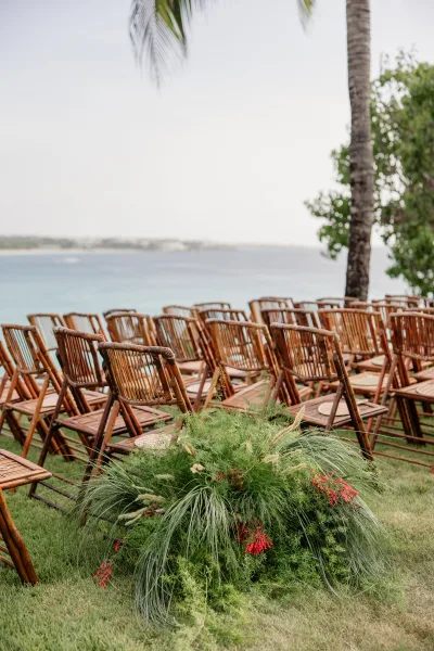 Ceremony seating features bamboo ceremony chairs along a grass lawn, facing the ocean with palm trees and red florals at the aisle.
