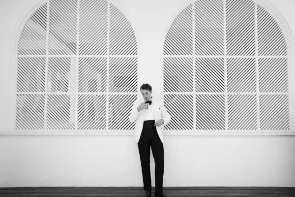 Groom portrait of a black tie groom in a white dinner jacket, posed by arched windows against striped panels and a white wall.