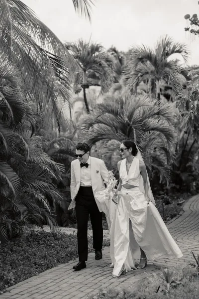 Couple portrait of bride and groom walking along a garden path lined with palm trees, both in sunglasses, amid tropical foliage under a cloudy sky.