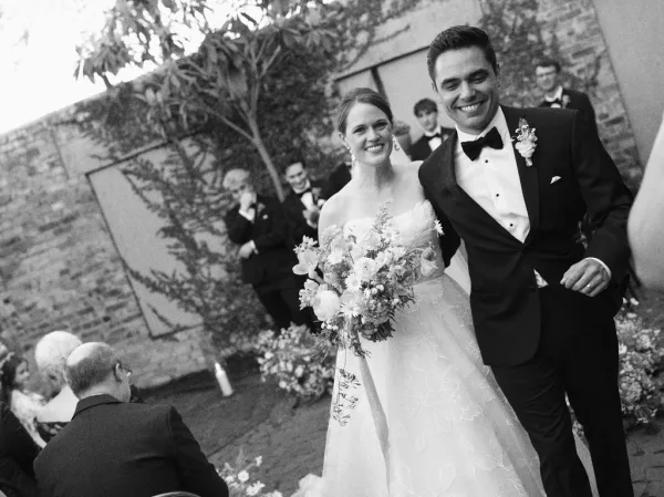 Ceremony recessional as a couple walking down the aisle, bride holding a bridal bouquet in a brick courtyard with garden foliage.