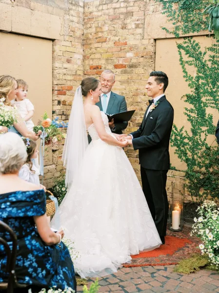 Wedding vows in a tender ceremony moment as the bride and groom hold hands by an ivy-covered brick wall in a courtyard, atop a vintage rug.