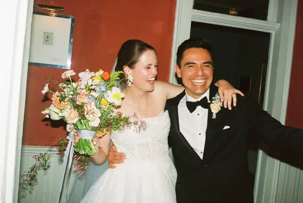 Couple portrait capturing a candid wedding moment as the bride and groom laugh, her bouquet held between them while they stand arm in arm.