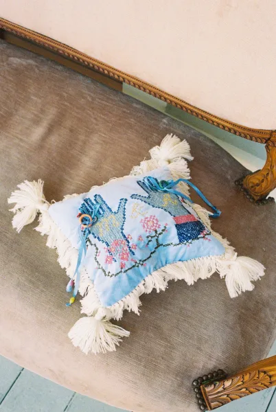 Ring bearer pillow serving as a wedding ring holder, detailed with floral embroidery, resting on a velvet chair with a carved wood arm.