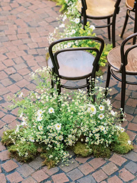 Ceremony aisle design with softly layered aisle flowers of white daisies edging the path across a cobblestone courtyard.