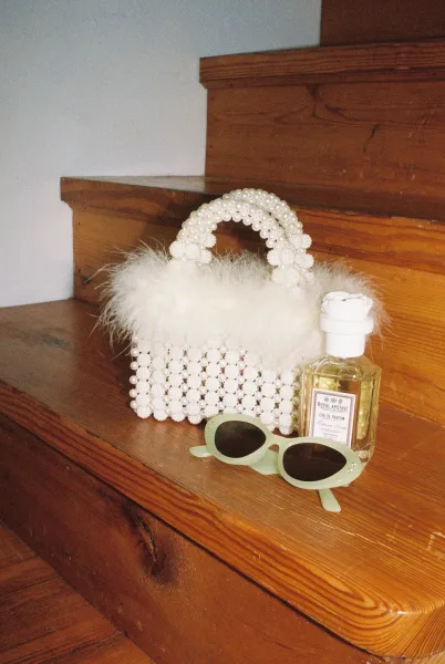 Bridal handbag, a pearl beaded bridal bag, rests on wood stairs beside sunglasses against a white wall.