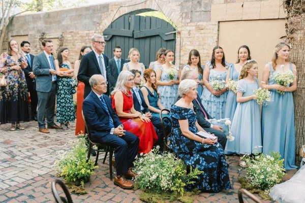 Ceremony moment at an outdoor courtyard ceremony as bridesmaids in blue line up on the brick courtyard by a rustic wooden gate and tree