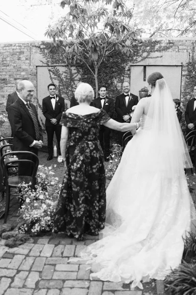 Wedding processional with a bride walking down the aisle, escorted by her mother, long tulle veil trailing past an ivy-clad brick wall in a courtyard.