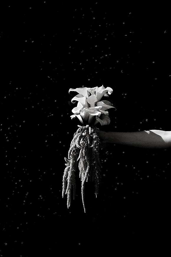 Bridal bouquet of white calla lilies with cascading greenery held on the bride’s arm against a dark backdrop as rain falls