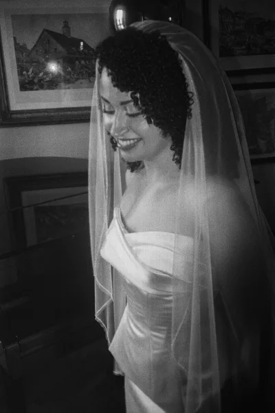 Bridal portrait in black and white of a smiling bride in a strapless satin wedding dress with veil over her face by a wood-paneled wall