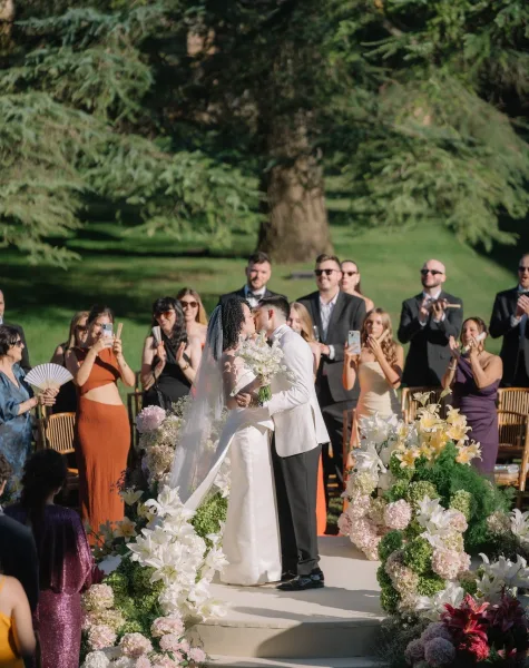 Ceremony kiss as bride in veil and groom in white dinner jacket embrace on a floral-lined aisle with guests on phones in a garden setting