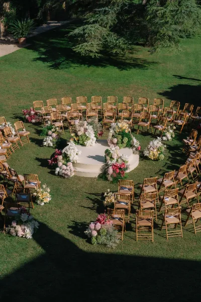 Ceremony setup with outdoor wedding ceremony seating around a round platform, wooden chairs with programs, and floral clusters on a shaded lawn