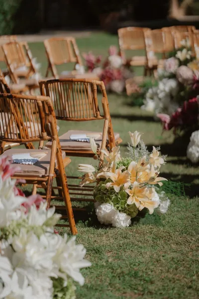 Ceremony aisle decor with outdoor ceremony chairs, rattan seating lined with lily and hydrangea floral clusters on a garden lawn