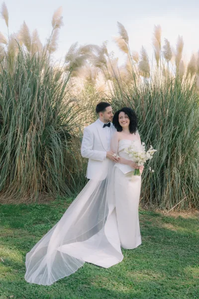 Couple portrait of bride and groom hugging, her strapless dress and calla lily bouquet framed by tall ornamental grass and sky