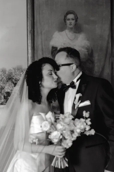 Wedding kiss portrait of bride and groom kissing, her veil and rose bouquet beside his tuxedo and glasses, framed painting behind them
