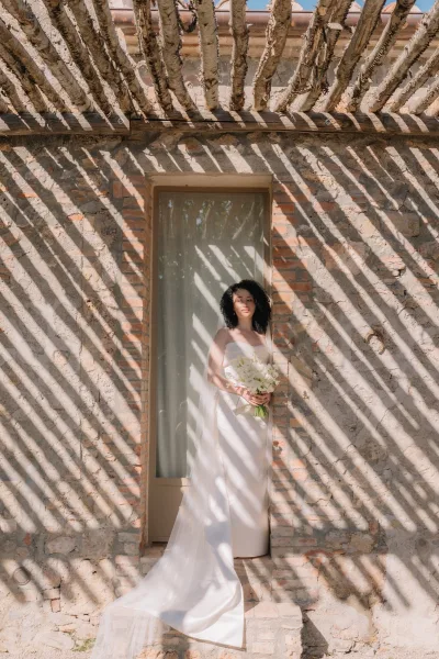 Bridal portrait of a bride holding bouquet, in a strapless satin gown with cathedral veil and white calla lilies by a sunlit stone wall doorway