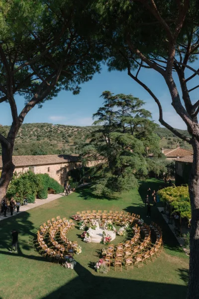 Outdoor ceremony setup with circular wedding ceremony seating around a white round platform, floral arrangements on a lawn by stone buildings under blue sky