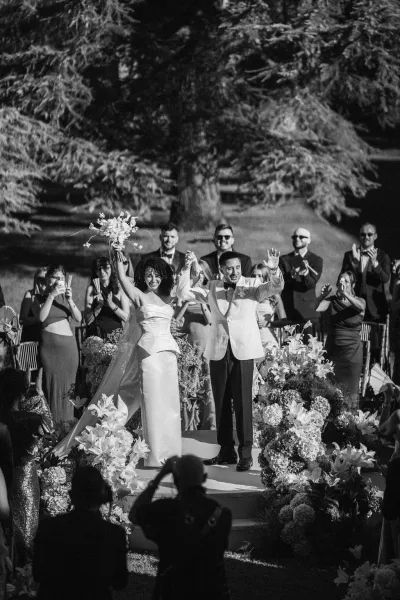 Wedding recessional as bride lifts bouquet overhead beside groom in tuxedo, walking a floral-lined aisle while guests cheer in a garden